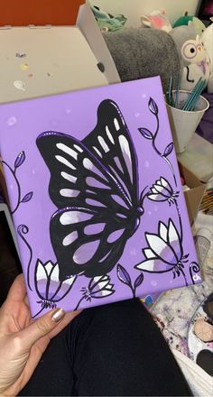 a woman holding up a purple and black butterfly painted on a canvas with white flowers