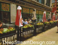 people sitting at tables in front of a restaurant with umbrellas and flower boxes on the sidewalk