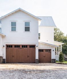 two brown garage doors are open in front of a white house with brick driveway and trees