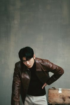 a young man leaning against a wooden box with his hands on his hips, wearing a brown leather jacket