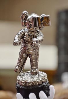 a silver statue of an astronaut standing on top of a black object with white hands