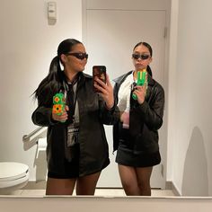 two women are taking pictures in the mirror with their cell phones and one is wearing sunglasses