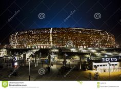 an illuminated stadium at night with lots of lights on the roof and people walking around