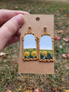 a pair of sunflowers painted on the side of an oval mirror earrings are held in front of a cardboard card