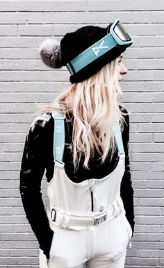 a woman with long blonde hair wearing white overalls and a black hat is standing in front of a brick wall