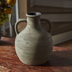 a gray vase sitting on top of a wooden table next to a bunch of grapes