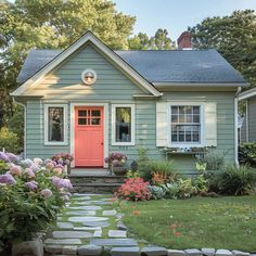 a small blue house with red door surrounded by flowers and greenery in the front yard