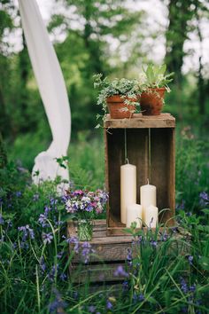 two white candles are sitting on a wooden crate in the middle of some purple flowers