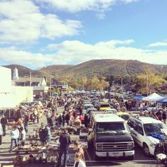 a crowd of people walking around a parking lot next to parked cars and tents with mountains in the background