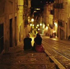 two people sitting on the ground next to each other in an alleyway at night