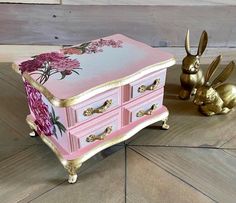 a pink and gold jewelry box sitting on top of a wooden floor next to two golden bunny figurines