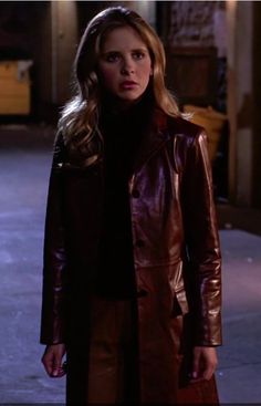 a woman wearing a brown leather coat and black turtle neck sweater standing in an alley