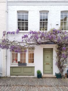 a white building with purple flowers growing on it's side and windows above the door