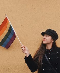 a woman holding a rainbow flag in front of a tan wall with a black hat