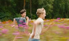 two young men running through a field full of purple and pink flowers with trees in the background