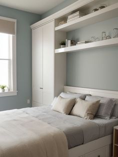 a white bed sitting under a window next to a shelf filled with dishes and cups