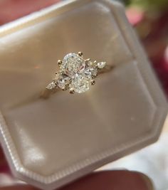 a diamond ring is being held in a white box with pink flowers behind it and someone's hand holding the ring