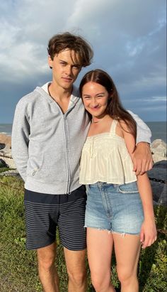 a man and woman standing next to each other on the grass by the ocean with their arms around each other