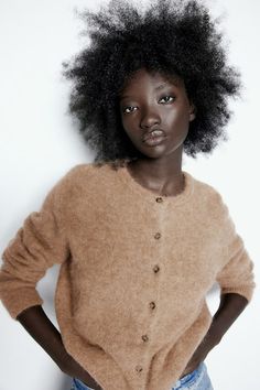 a woman with an afro standing in front of a white wall wearing a brown cardigan