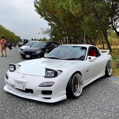 a white sports car parked on the side of a road next to some trees and people