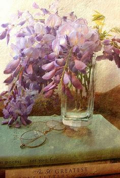 a painting of purple flowers in a glass vase on top of a book with glasses