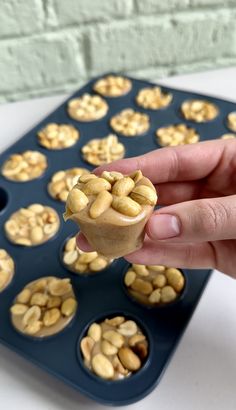 a hand holding a cupcake in front of a muffin tin filled with nuts