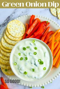 Green onion dip in a white bowl served with crackers and veggie sticks.