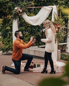 a man kneeling down next to a woman in front of a white drape and flowers