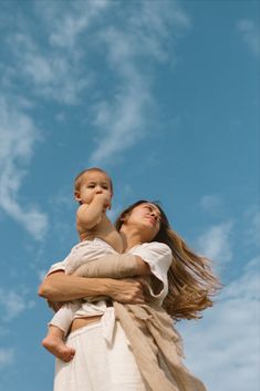 a woman holding a baby up in the air against a blue sky with wispy clouds