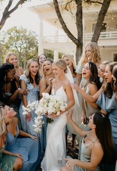 a group of women standing around each other in front of a white house and holding bouquets