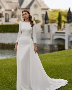 a woman in a long white dress standing on the grass near a building and water