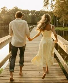 a man and woman holding hands while walking on a wooden bridge over a lake at sunset