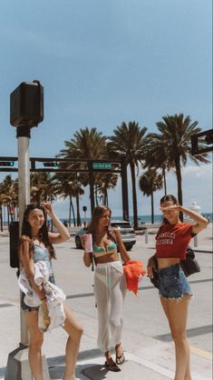 three young women standing on the sidewalk near a street sign and palm trees in the background