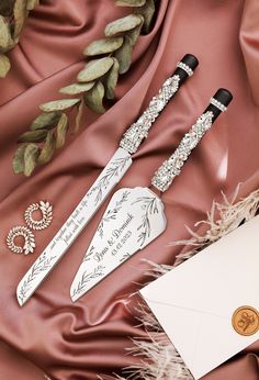 two decorative knifes are laying on a red satin surface next to an envelope and card