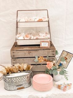 an assortment of items displayed on a table with white linens and pink plates in metal containers