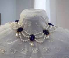 In the late 1790s, a style of necklace called a "festoon" came in to fashion. It is a lovely and distinctive style that features swags of pearl or chain that come together in central focal points. It's one of my favorite styles of jewelry! They remained popular through the Edwardian Era. This particular version is made with small white glass pearls and is linked with Glass Lapis stones in a rich, shining blue. They are set in crown settings and a teardrop dangle pearl hanging from each. The cent Baroque Necklaces With Historical Design For Wedding, Baroque Necklace With Historical Design For Weddings, Elegant Baroque Necklace With Historical Design, Baroque Wedding Necklace With Historical Design, Baroque Historical Design Necklace For Wedding, Elegant Pendant Necklaces With Historical Design, Elegant Historical Pendant Necklaces, Victorian Pearl Drop Necklace, Victorian Pearl Necklace For Formal Occasions