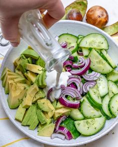 someone pouring dressing into a bowl filled with cucumber, onions and avocado