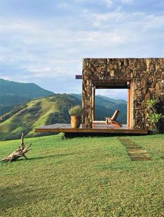 a house made out of rocks sitting on top of a lush green hillside with mountains in the background