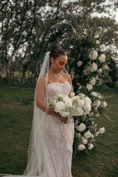 a woman in a wedding dress standing next to a white flower covered arch with greenery