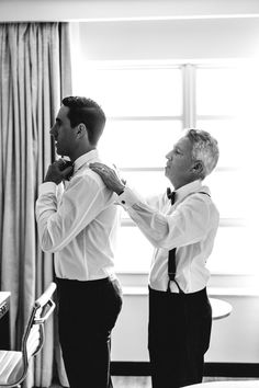 two men standing next to each other in front of a window tying their ties together