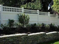 a white fence and some plants in front of a stone wall with bushes on the other side