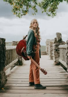 a woman standing on a bridge with a guitar in her hand and wearing overalls