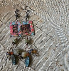 The Tropics Earrings, so named for their bright, tropical colors of coral and green, feature enameled copper charms by TendancePerles with a cut-out design bordered by imagery of exotic flora. Square-headed eye pins are above and below, the latter supporting dangles of tourmaline, coral, green garnet, contemporary Czech glass, and brass heishi. From attachment to ear wire they measure about 4 1/4 inches, with dangles measuring about 1 inch. Each earring weighs .3 ounce or 10 grams. They are on hypoallergenic Niobium ear wires. Coral And Green, Green Garnet, Enameled Copper, Tropical Colors, Eye Pins, Cut Out Design, Ear Wire, Czech Glass, Earrings Handmade