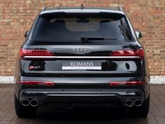 the rear end of a black car parked in front of a brick wall with an inscription on it