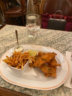 a white plate topped with fish and fries next to a glass of wine on top of a table