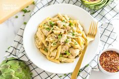 a white bowl filled with pasta next to an artichoke and some seasoning