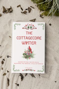 a book sitting on top of a sandy beach next to grass and leaves, with the title'the cottage core winter'written in red