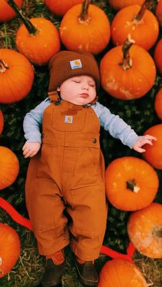 a baby laying on top of a pile of pumpkins wearing overalls and a hat