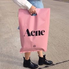 a woman holding a pink bag with the word acne printed on it and black boots