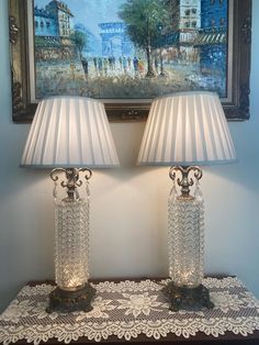 two glass lamps sitting on top of a table next to a wall with a painting above it